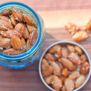 An overhead view of a blue jar of maple coconut roasted almonds. There is a small dish next to the jar with additional almonds in it.