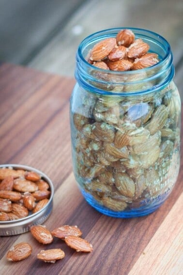 A blue mason jar filled to the top with maple coconut roasted almonds. The lid is laying next to the jar with additional almonds in it and a few scattered in front of it.