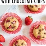 Strawberry protein muffins with chocolate chips in red polka dot cupcake liners on a table with strawberries around them.