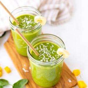 Two Ball mason jars filled with a green smoothie sitting on a wooden cutting board with spinach, mango and coconut shreds scattered around.