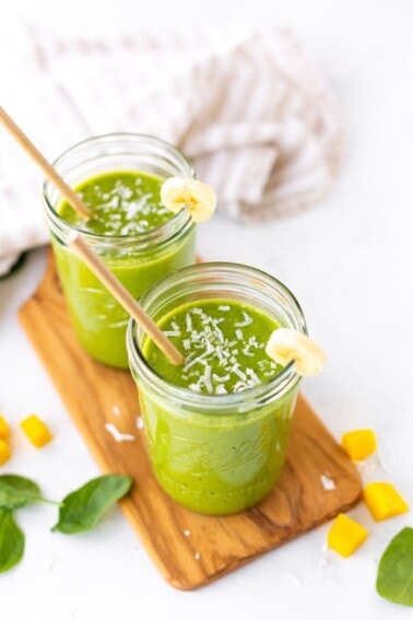 Two Ball mason jars filled with a green smoothie sitting on a wooden cutting board with spinach, mango and coconut shreds scattered around.