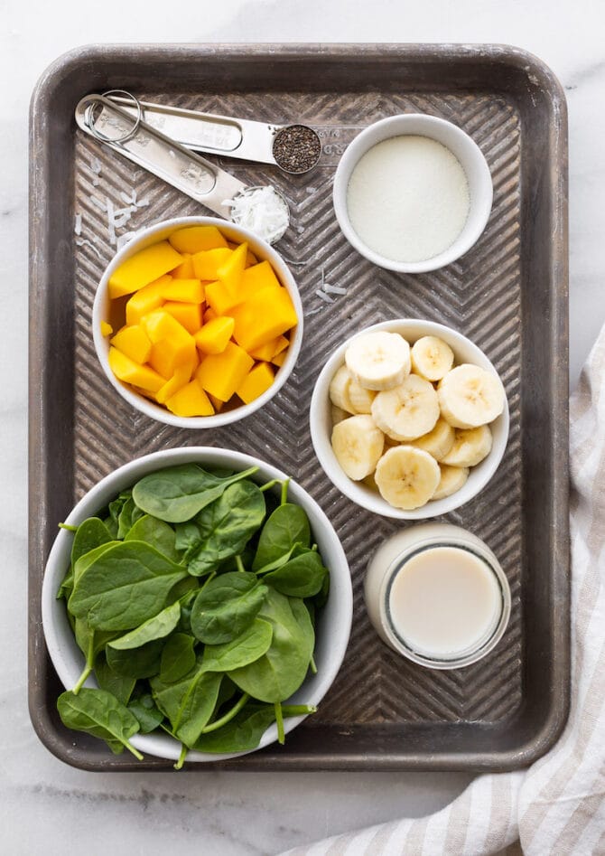 Ingredients for tropical green smoothie laid out on a baking tray: baby spinach, milk, banana slices, mango chunks, protein powder, chia seeds and shredded coconut.