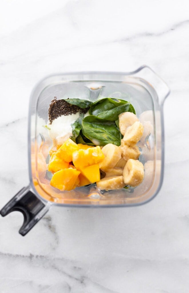 A blender container filled with mango, banana, spinach, coconut, chia seeds and almond milk