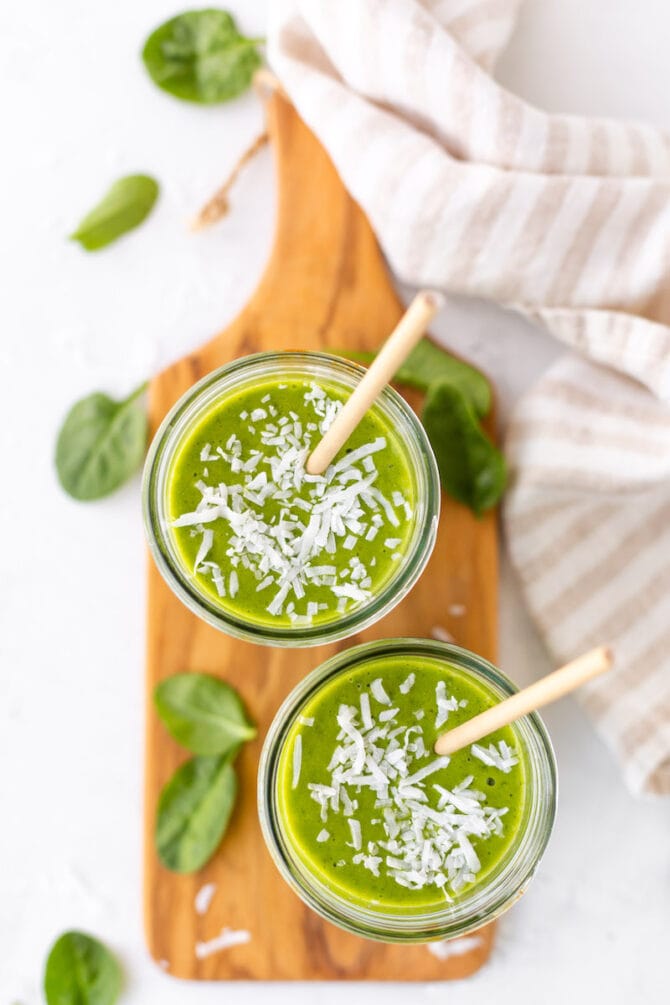 Overhead view of two Ball mason jars filled with a green smoothie sitting on a wooden cutting board with spinach scattered around.