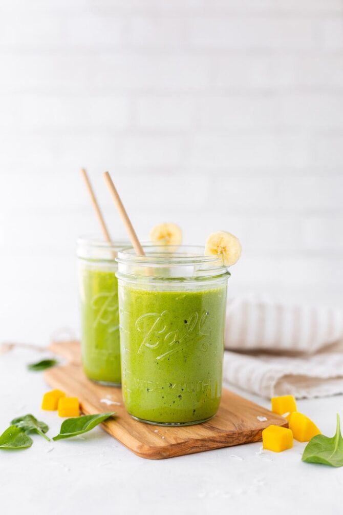 Two Ball mason jars filled with a green smoothie sitting on a wooden cutting board with mango chunks and spinach scattered around,