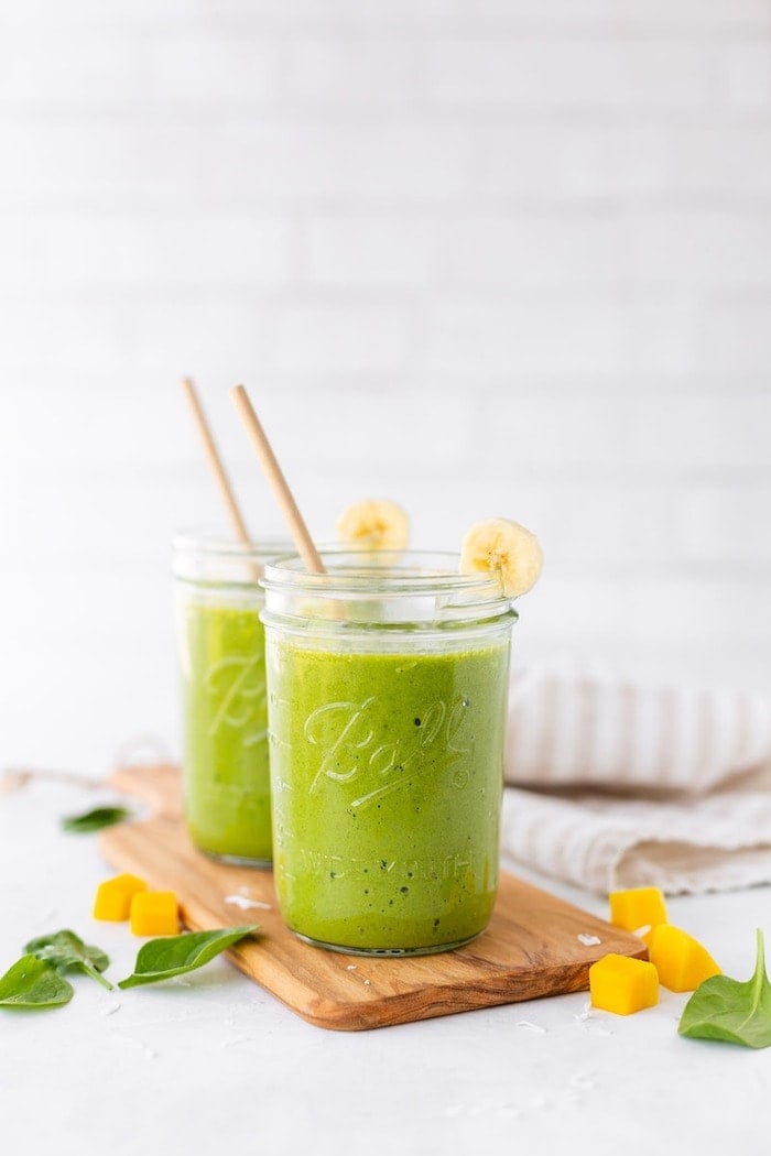 Two Ball mason jars filled with a green smoothie, garnished with a banana slice, sitting on a wooden cutting board with mango chunks and spinach scattered around.