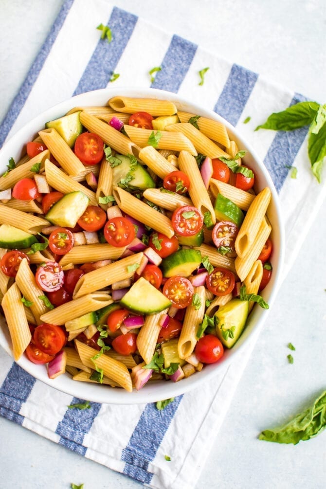 Bowl of penne pasta salad with basil, cucumbers, tomatoes and red onions. Bowl is on a striped kitchen towel.