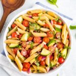 Bowl of clean eating penne pasta salad mixed with basil, cherry tomatoes, cucumber and red onion. Next to the bowl are fresh basil leaves and a wooden mixing spoon.