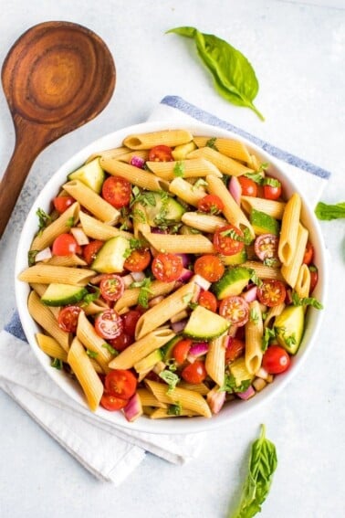 Bowl of clean eating penne pasta salad mixed with basil, cherry tomatoes, cucumber and red onion. Next to the bowl are fresh basil leaves and a wooden mixing spoon.