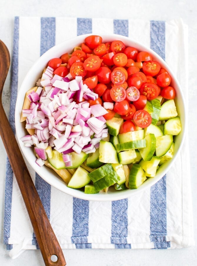 Bowl with ingredients for healthy pasta salad before mixing. In the bowl, on top fo penne are sections of chopped red onion, cherry tomatoes and chopped cucumber. Wood spoon and striped dish towel are by the bowl.