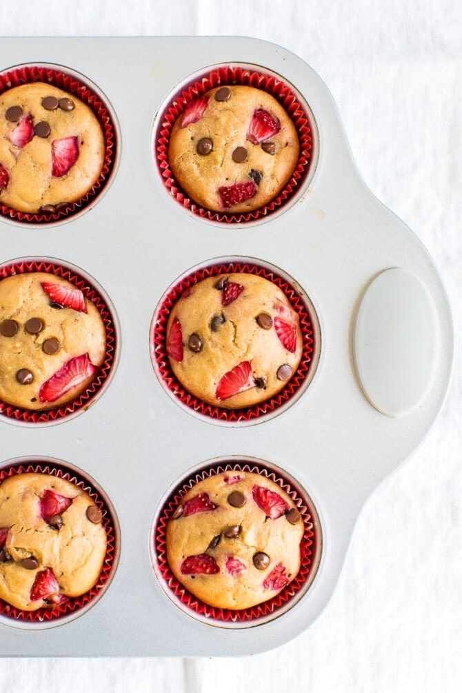Strawberry chocolate chip protein muffins in red cupcake liners in a muffin tin. Muffins are topped with chopped strawberries and mini chocolate chips.