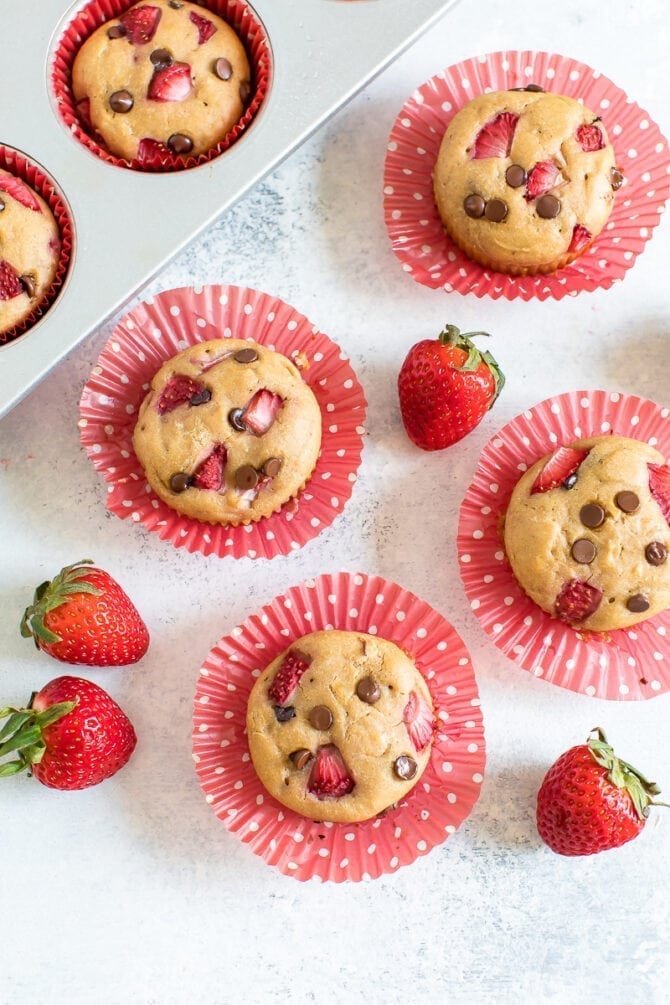 Strawberry protein muffins with chocolate chips in red polka dot cupcake liners on a table with strawberries around them.