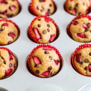 Strawberry chocolate chip protein muffins in red cupcake liners in a muffin tin. Muffins are topped with chopped strawberries and mini chocolate chips.