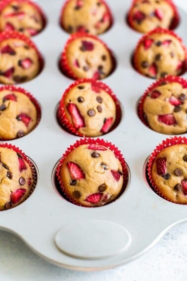 Strawberry chocolate chip protein muffins in red cupcake liners in a muffin tin. Muffins are topped with chopped strawberries and mini chocolate chips.