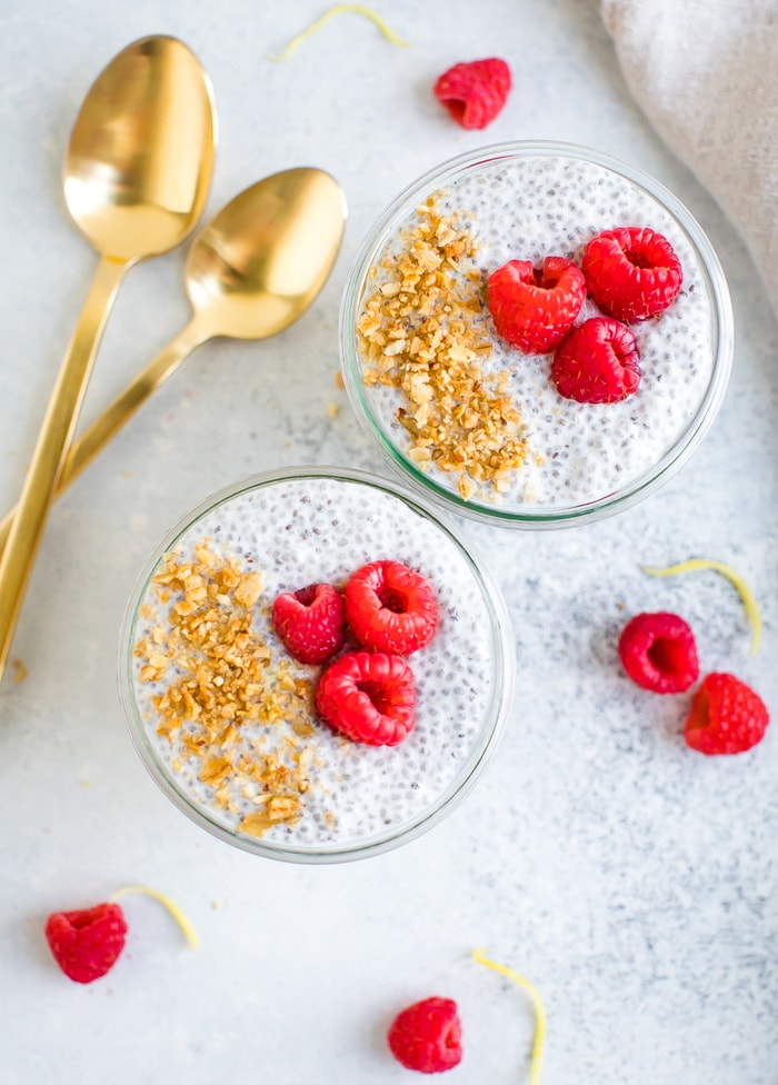 Overhead photo of two jars of raspberry chia pudding topped with ground granola and raspberries. Spoons and berries are around the jars on the table.