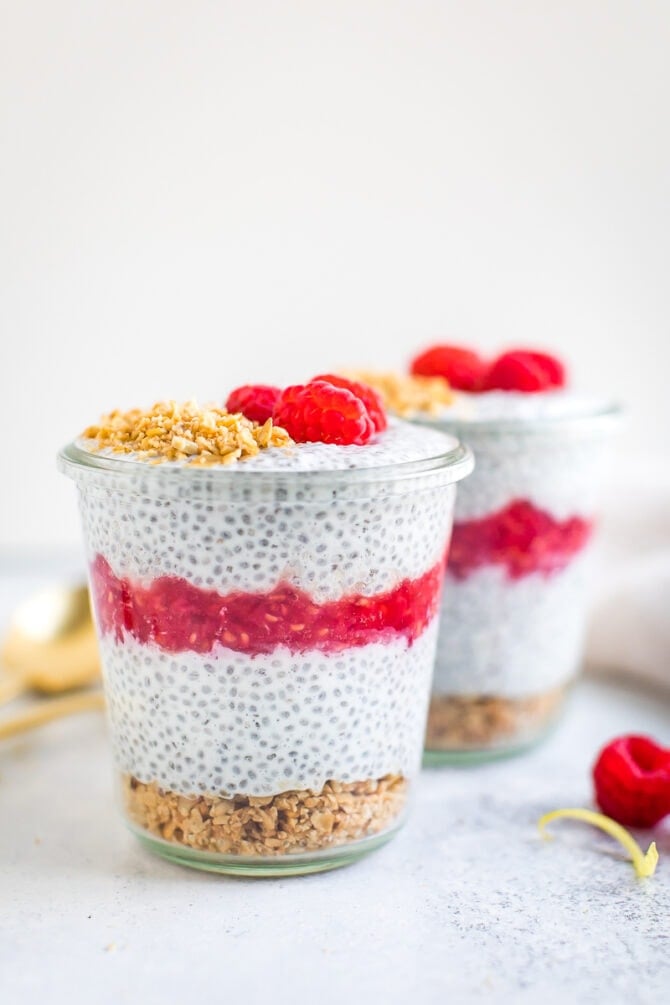 Two jars of chia pudding layered with raspberry puree and topped with granola and raspberries.