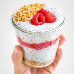 Hand holding a jar of raspberry cheesecake chis pudding layered with raspberries and topped with granola and raspberries.