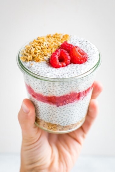 Hand holding a jar of raspberry cheesecake chis pudding layered with raspberries and topped with granola and raspberries.