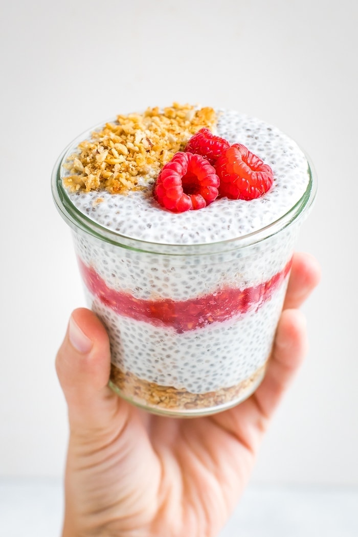 Hand holding a jar of raspberry cheesecake chis pudding layered with raspberries and topped with granola and raspberries.