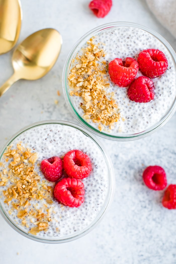Overhead photo of two jars of raspberry chia pudding topped with ground granola and raspberries. Spoons and berries are around the jars on the table.