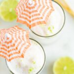 Overhead shot with two skinny coconut daiquiri cocktails in tall glasses with orange and white striped cocktail umbrellas.