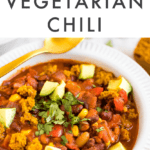Easy vegetarian chili in a bowl, topped with cilantro, avocado, and bits of cornbread muffin. A spoon, cilantro and cornbread muffin are laying around the healthy bowl of chili.