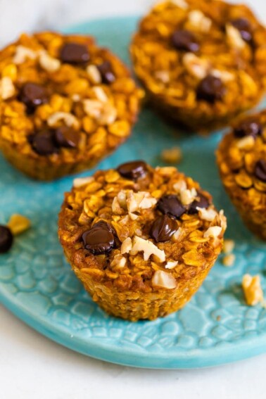 Four baked pumpkin oatmeal cups on a plate and one on the table next to the plate. Oatmeal cups are topped with chocolate chips and walnuts.