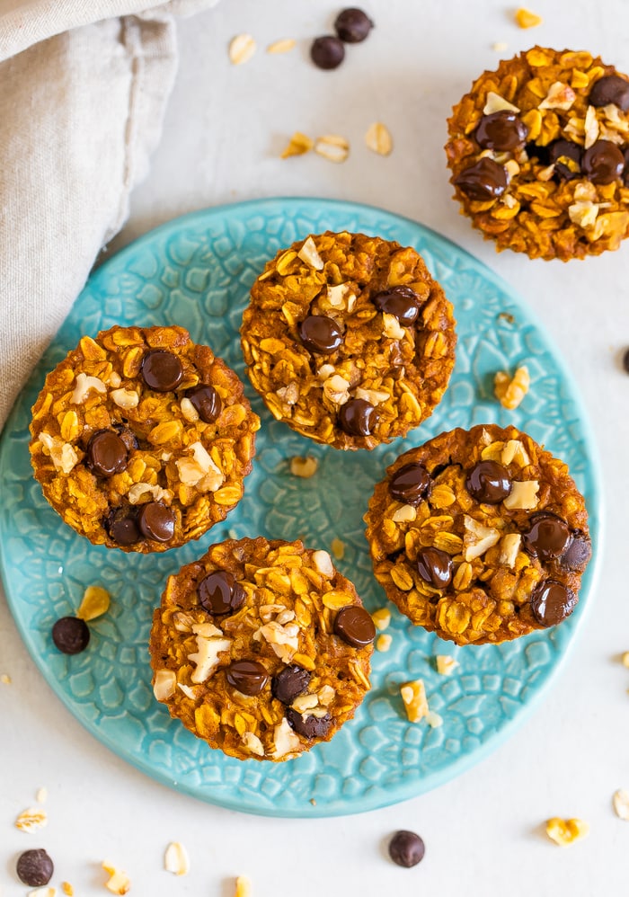 Four baked pumpkin oatmeal cups on a plate and one on the table next to the plate. Oatmeal cups are topped with chocolate chips and walnuts.