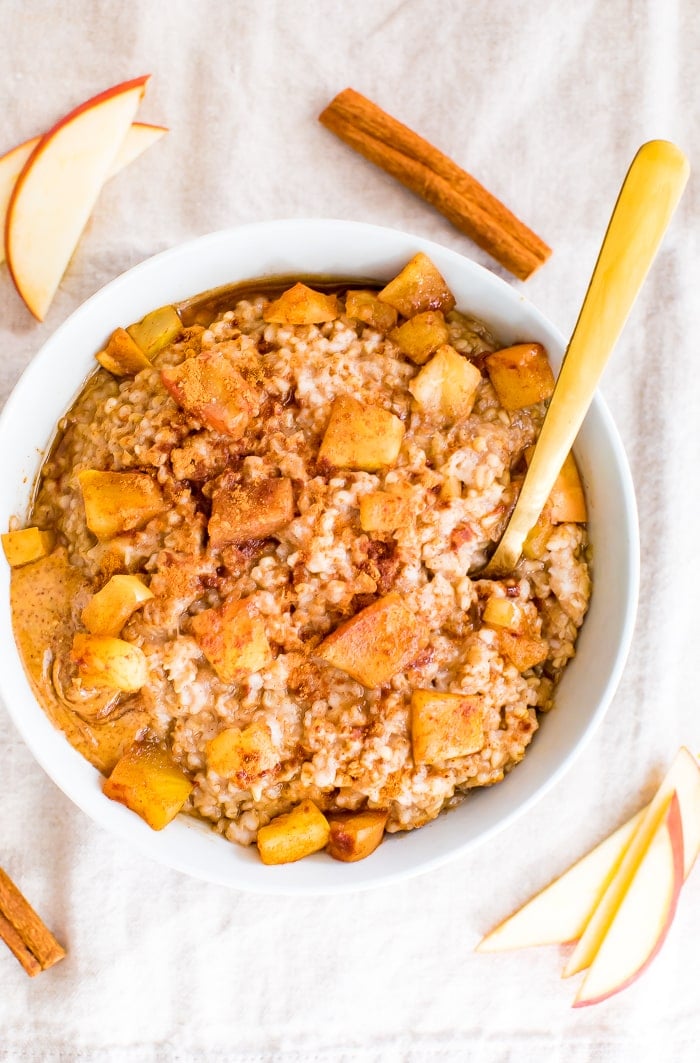 Bowl of healthy apple cinnamon oatmeal topped with chunks of apples, cinnamon, and peanut butter. Slices of apple and a stick of cinnamon are around the bowl.