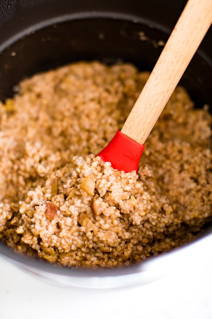 Spatula stirring a crock pot of healthy apple cinnamon oatmeal made with steel cut oats and apple chunks.