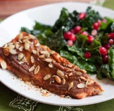 A filet of pumpkin spice salmon on a white plate next to a kale salad with pomegranate arils.