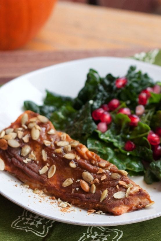 A filet of pumpkin spice salmon on a white plate next to a kale salad with pomegranate arils.