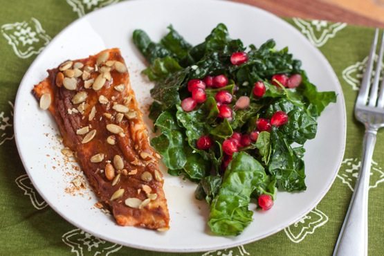 A filet of pumpkin spice salmon on a white plate next to a kale salad with pomegranate arils. A fork is laying next to the plate.