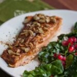 A filet of pumpkin spice salmon on a white plate next to a kale salad with pomegranate arils.