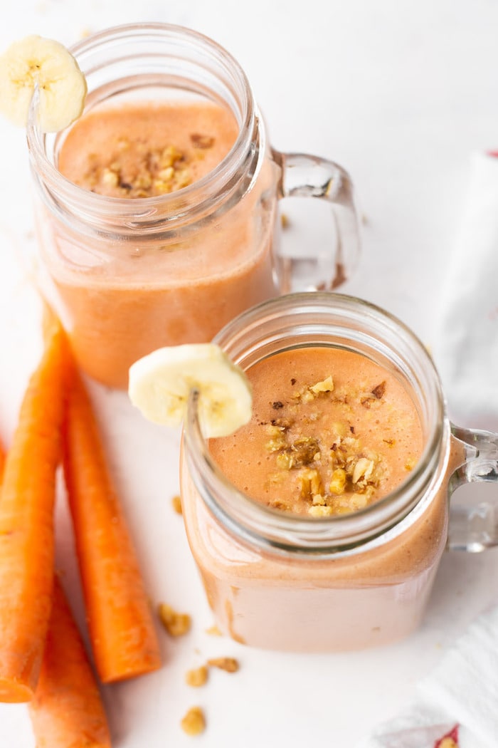 Two mason jar mugs full of carrot cake protein smoothies and topped with chopped walnuts and a banana slice on the rim.