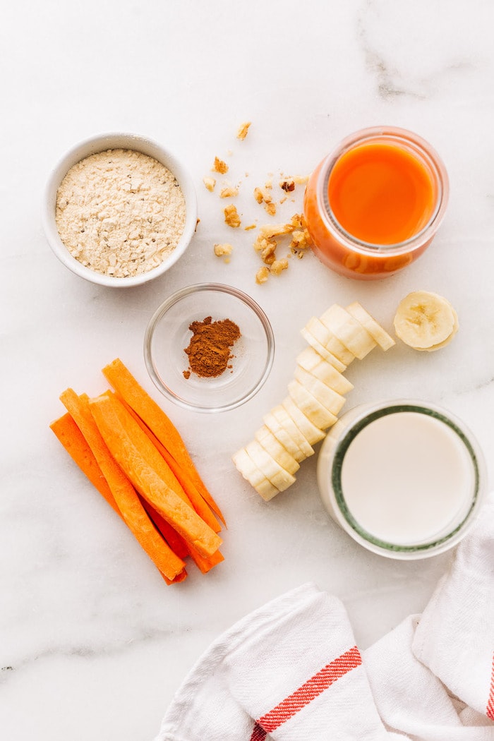 Ingredients to make carrot cake protein smoothie separated out on a table: carrot juice, protein powder, almond milk, cinnamon and sliced frozen banana.