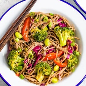 Bowl of soba noodle salad, with red pepper, broccoli, edamame, and purple cabbage, and topped with sesame seeds.