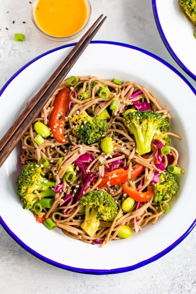 Bowl of soba noodle salad, with red pepper, broccoli, edamame, and purple cabbage, and topped with sesame seeds.