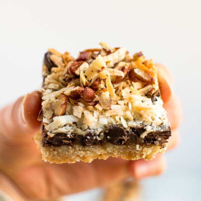 Hand holding a healthier, gluten free, grain free, vegan, magic cookie bar. An almond flour cookie crust topped with coconut caramel, coconut, pecans, and chocolate chips.