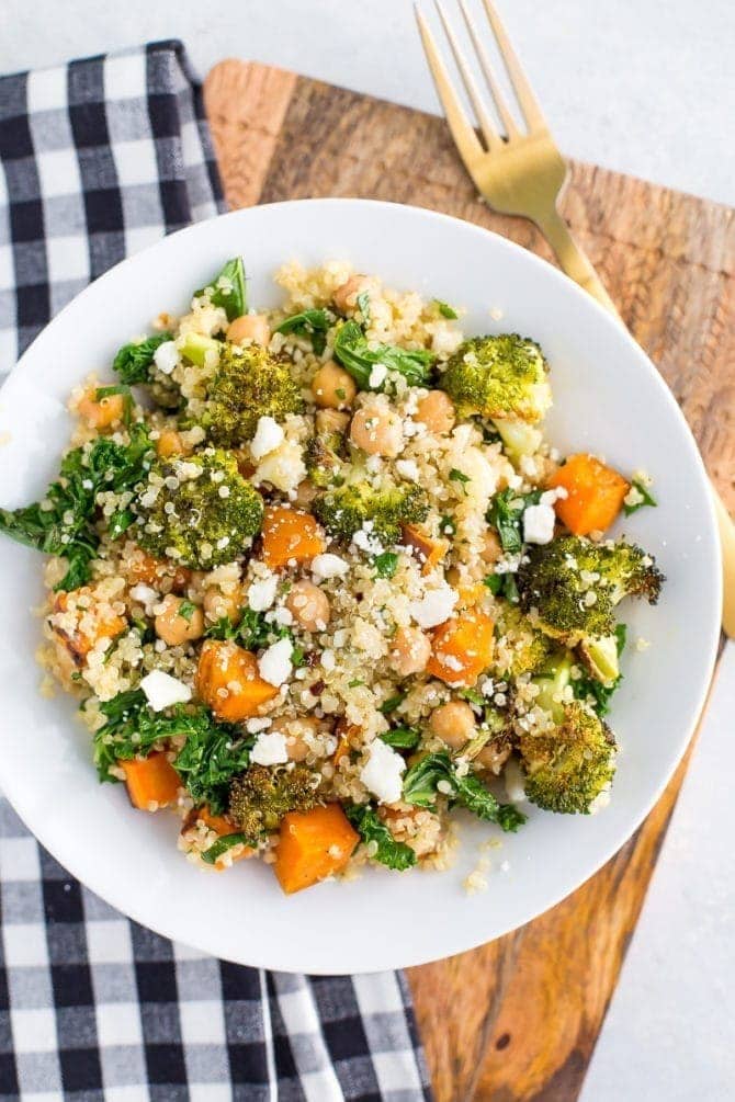 A white plate piled with roasted broccoli quinoa salad. Salad also has kale, sweet potatoes, feta, and chickpeas. Checkered napkin below the plate and a fork to the side.