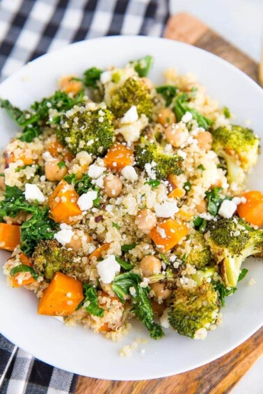 A white plate piled with roasted broccoli and quinoa salad. Salad also has kale, sweet potatoes, feta, and chickpeas. Checkered napkin below the plate.