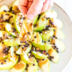 Hand holding an apple nacho drizzled with date caramel and sprinkled with mini chocolate chips and chopped pecans.