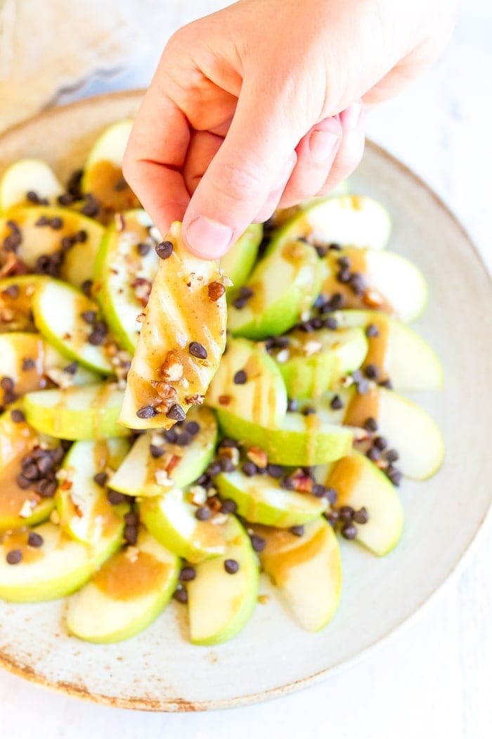 Hand holding an apple nacho drizzled with date caramel and sprinkled with mini chocolate chips and chopped pecans.