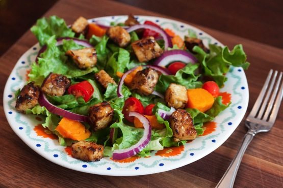 An overhead photo of a bed of salad greens with red onion slices and maple lemon tempeh cubes on a dinner plate. There is a silver fork laying next to the plate.