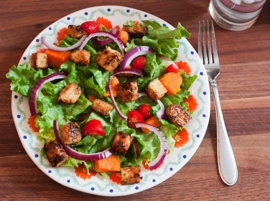 An overhead photo of a bed of salad greens with red onion slices and maple lemon tempeh cubes on a dinner plate. There is a silver fork laying next to the plate.