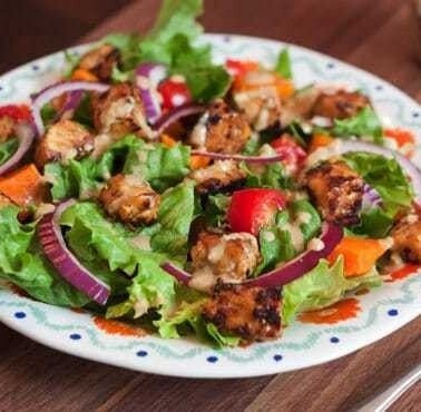 A bed of salad greens with red onion slices and maple lemon tempeh cubes on a dinner plate. There is a silver fork laying next to the plate.