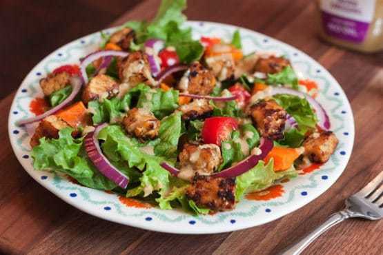 A bed of salad greens with red onion slices and maple lemon tempeh cubes on a dinner plate. There is a silver fork laying next to the plate.