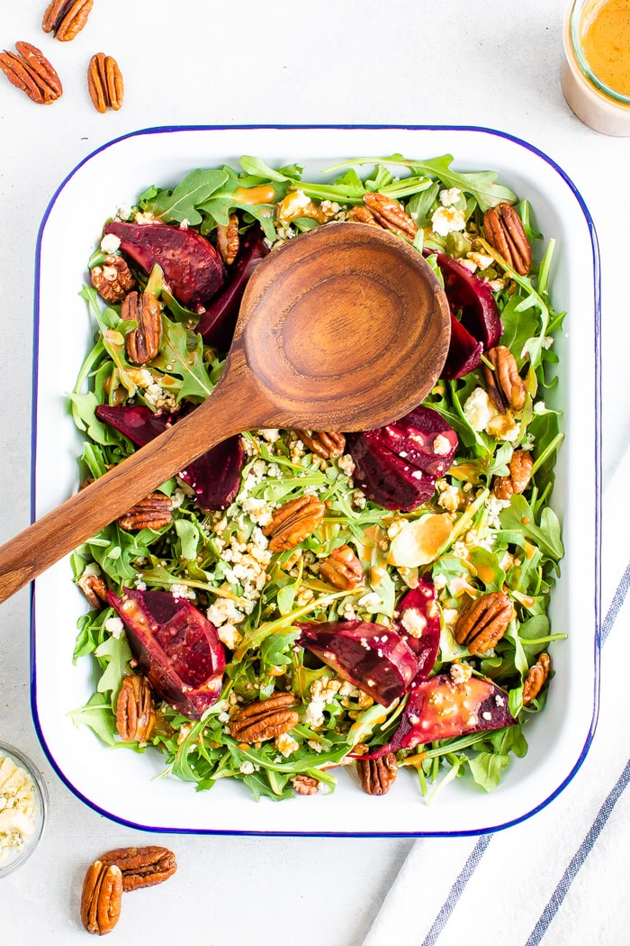 Rectangle serving tray and wooden spoon with arugula and beet salad topped with pecans and gorgonzola cheese.