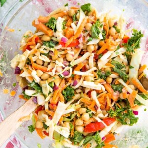 Glass bowl with wooden salad spoons tossing a salad made from kale, cabbage, chickpeas, carrots, onion, and peppers. Sprigs of cilantro are to the side on the table.