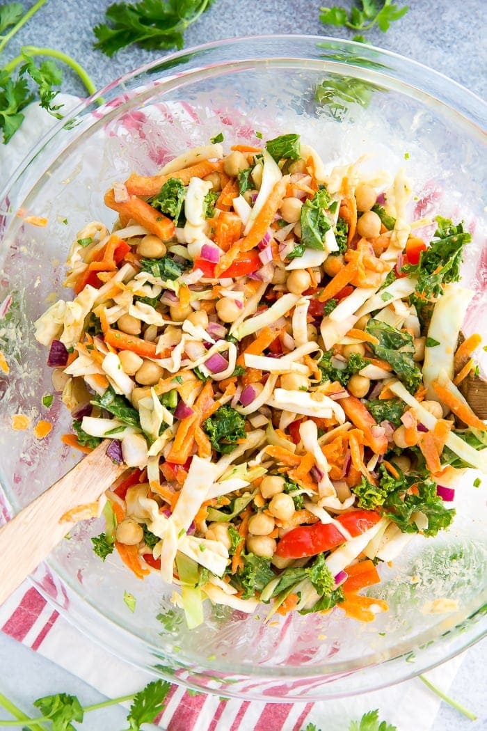Glass bowl with wooden salad spoons tossing a salad made from kale, cabbage, chickpeas, carrots, onion, and peppers. Sprigs of cilantro are to the side on the table.